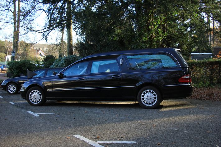 Una station wagon nera in stile carro funebre parcheggiata su una strada alberata accanto a un muro di pietra.