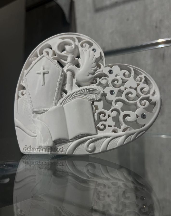 Cuore decorativo bianco con croce, libro aperto e volute in filigrana su una superficie riflettente.