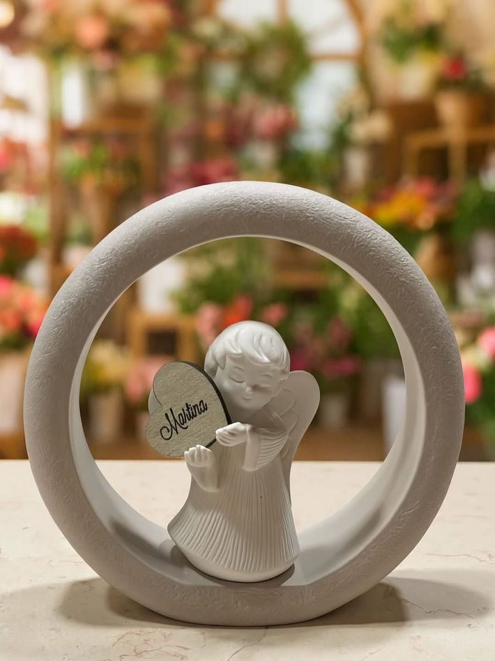 Anello in ceramica bianca con una piccola statuetta di un angelo che tiene in mano un cuore, su uno sfondo floreale sfocato.