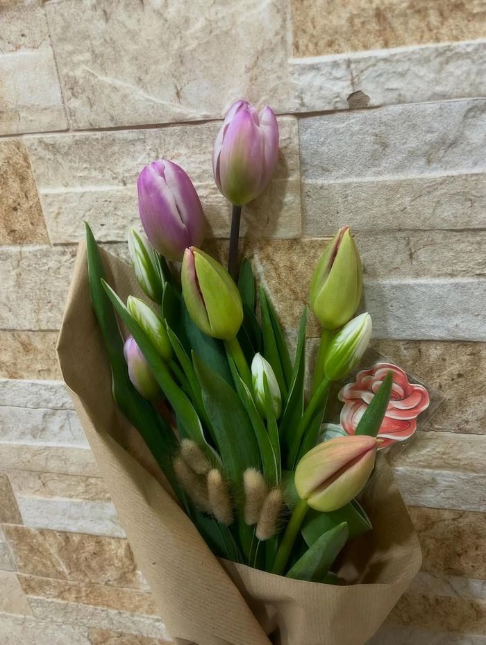 Bouquet di tulipani rosa e boccioli verdi avvolti in carta marrone, appoggiato a un muro di pietra.