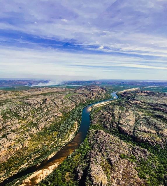 Aerial view of a winding river cutting through rocky green hills under a cloudy blue sky