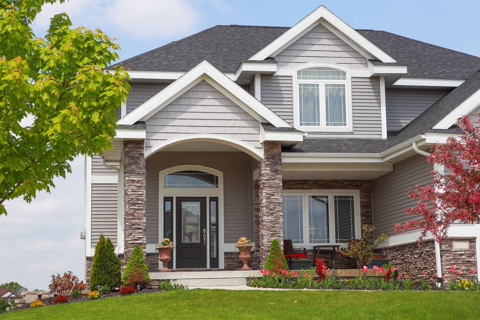 Two-story gray house with stone pillars, arched entryway, and landscaped front yard.