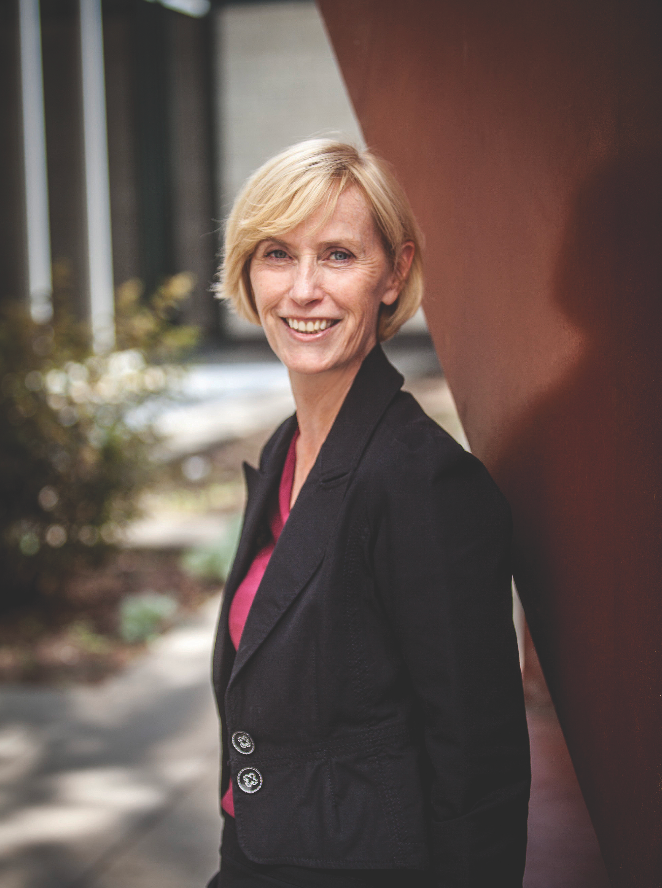 A woman in a black jacket is leaning against a wall and smiling.
