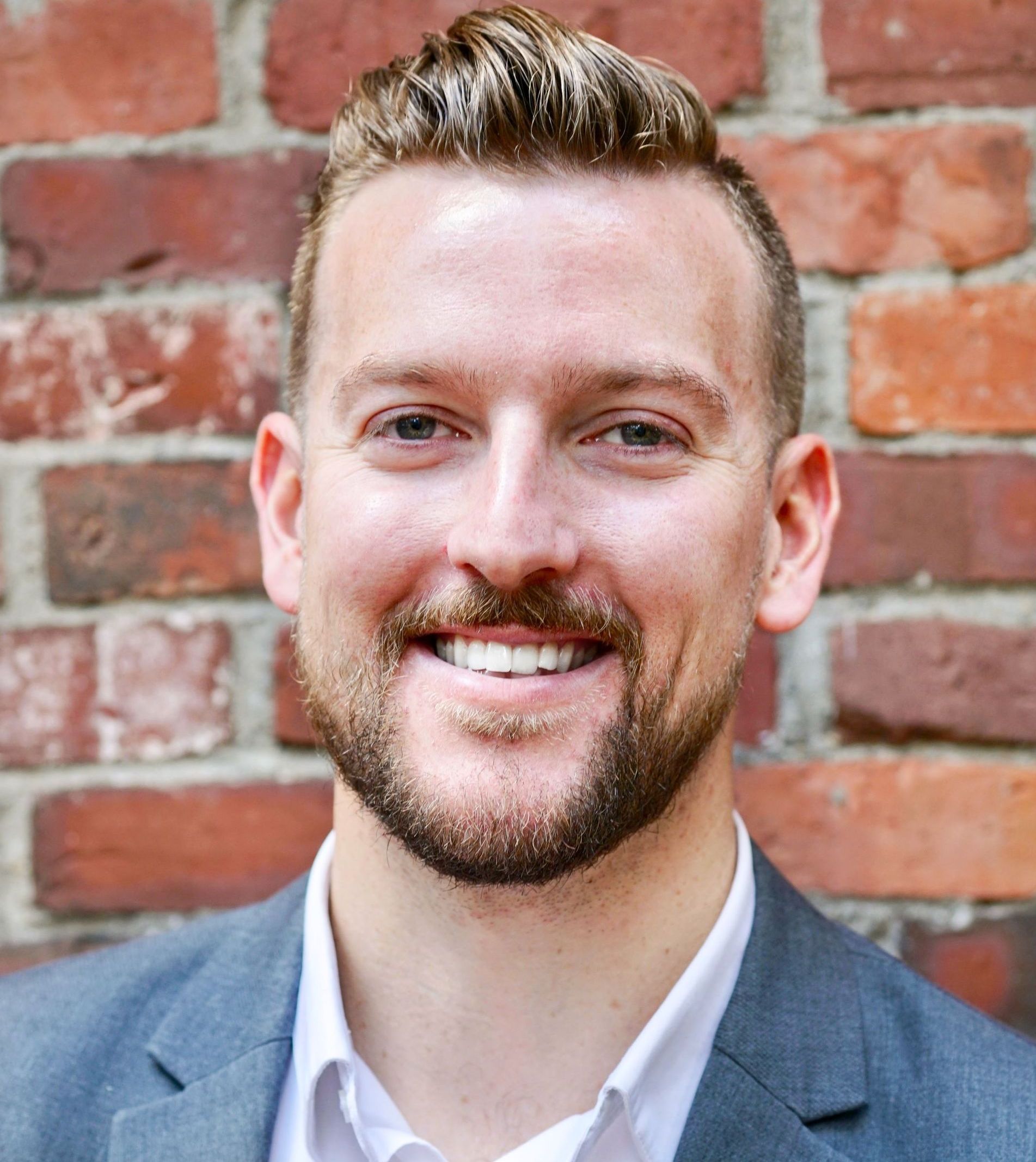 A man with a beard is smiling in front of a brick wall.