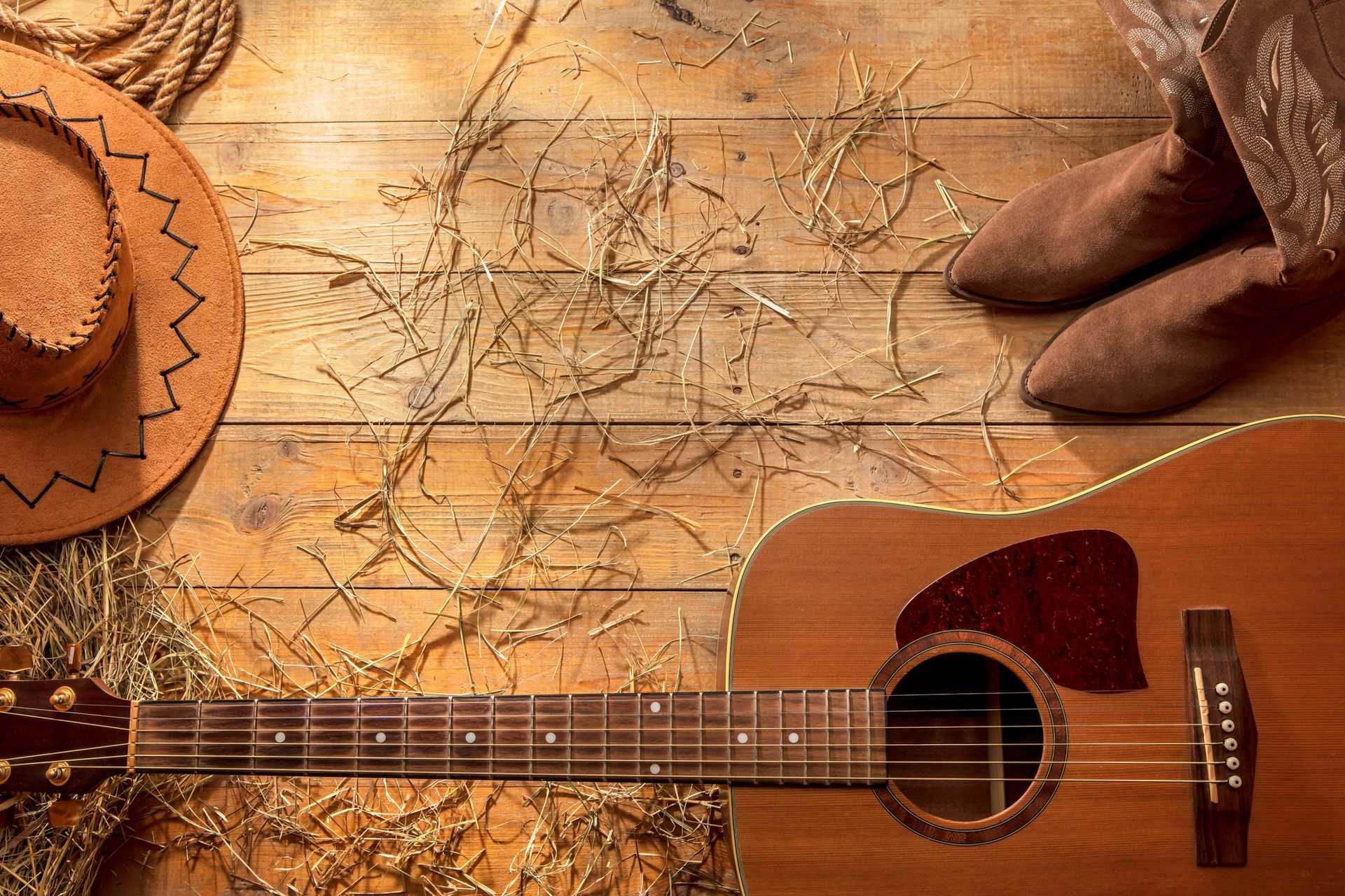 A guitar , cowboy hat , cowboy boots and hay are on a wooden table.