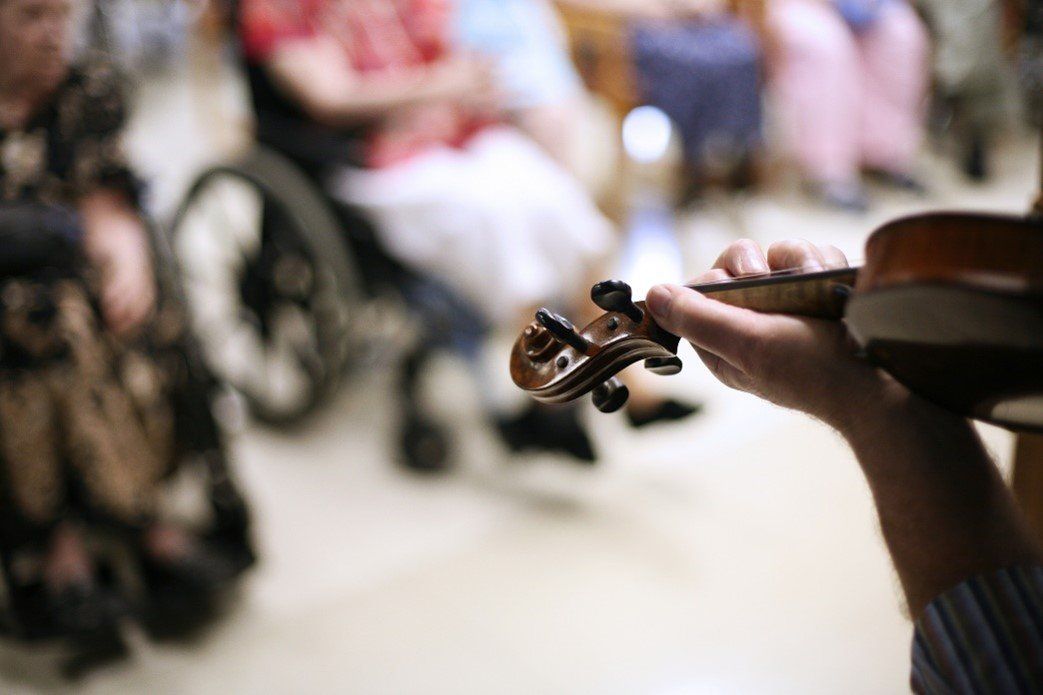 Close-up of hands holding a violin; people in a room, including one in a wheelchair, in the background.