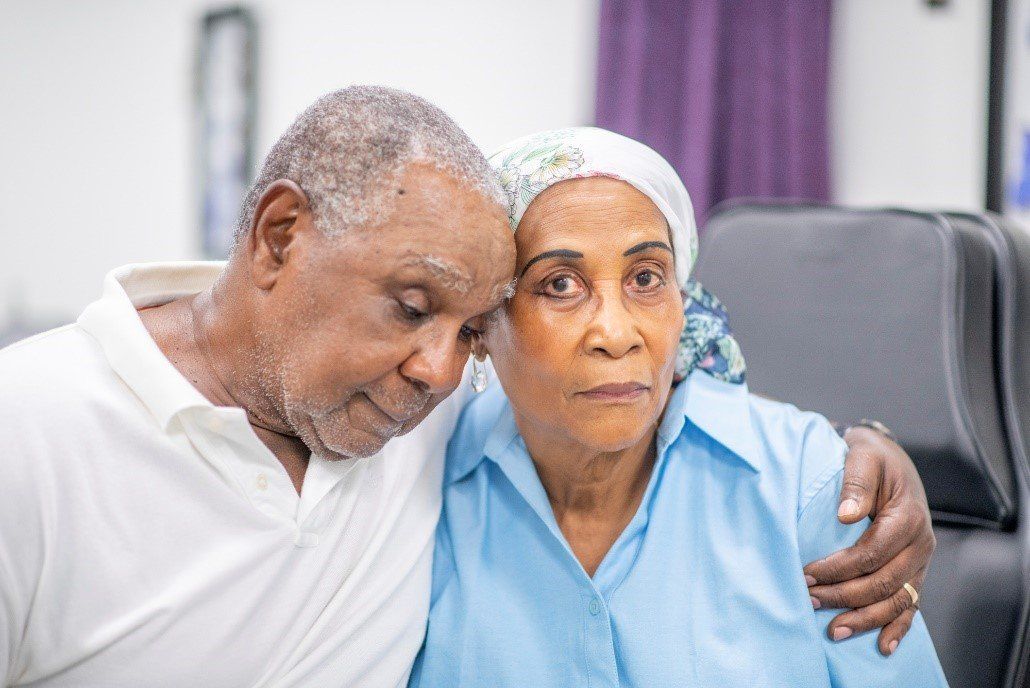 An older man in white shirt embraces a woman in blue shirt, head resting on her shoulder.
