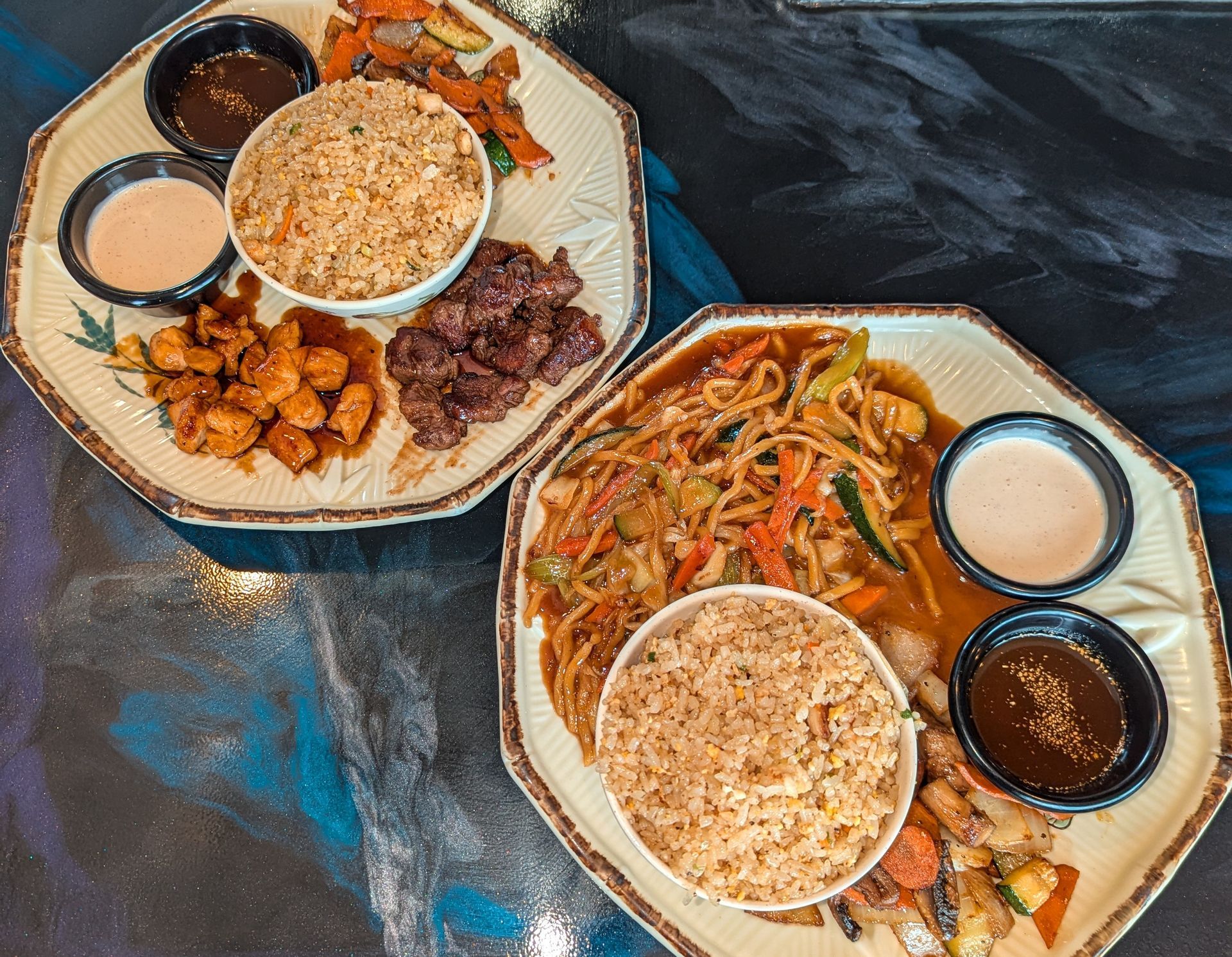 Two takeout meals on octagonal plates with rice, saucy dishes, and dipping cups on a dark table.