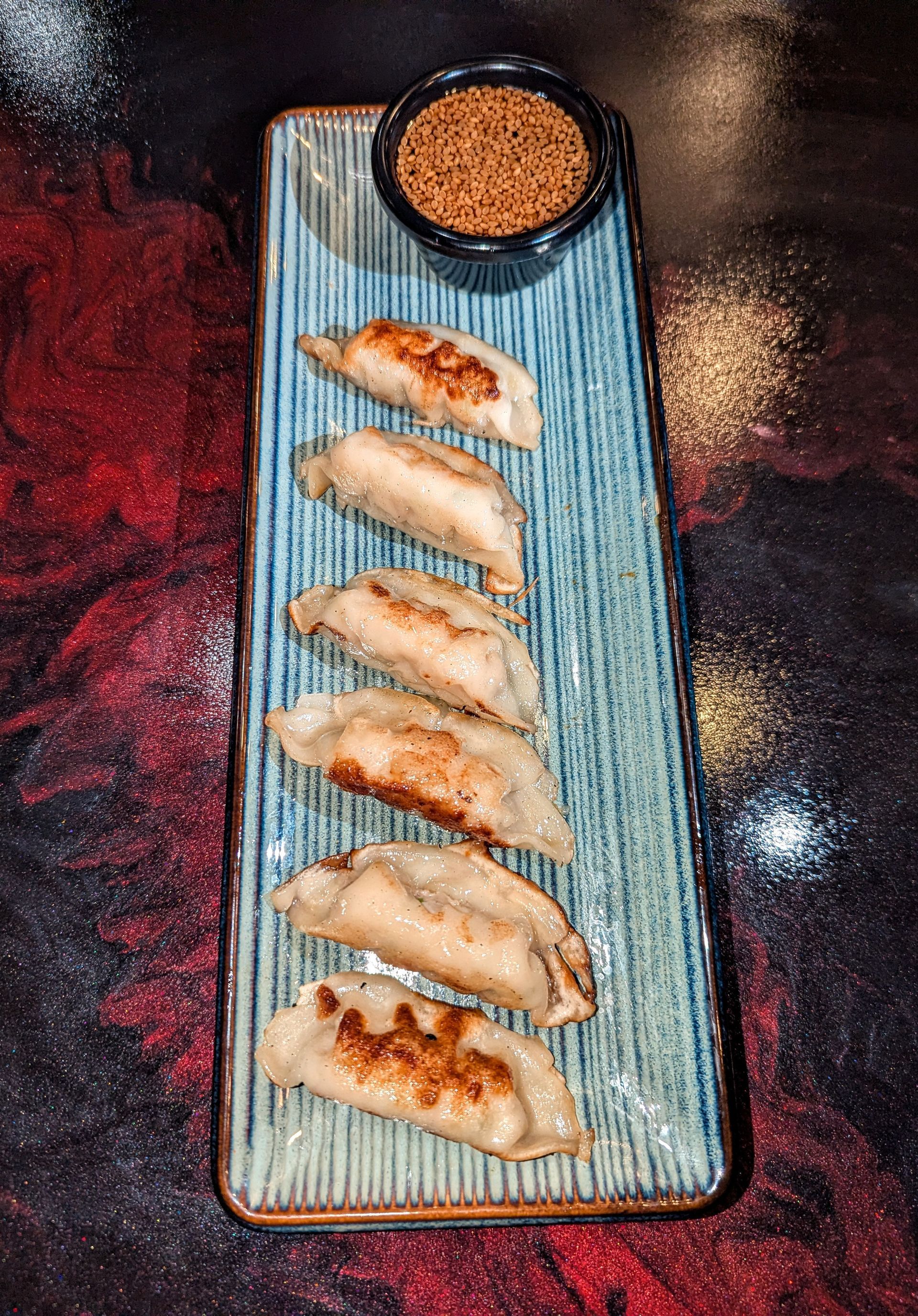 Five dumplings on a blue plate with a small bowl of dipping sauce on a dark table.