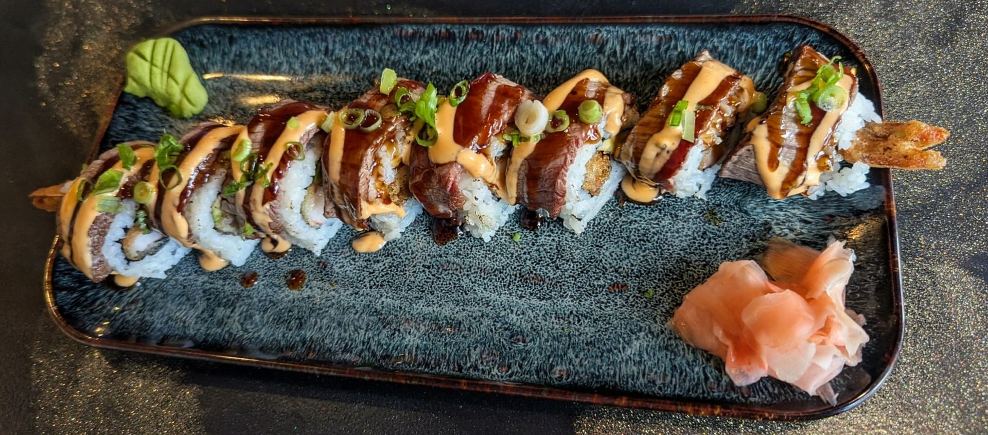 Sushi roll drizzled with sauce on a black plate, with wasabi and pickled ginger.