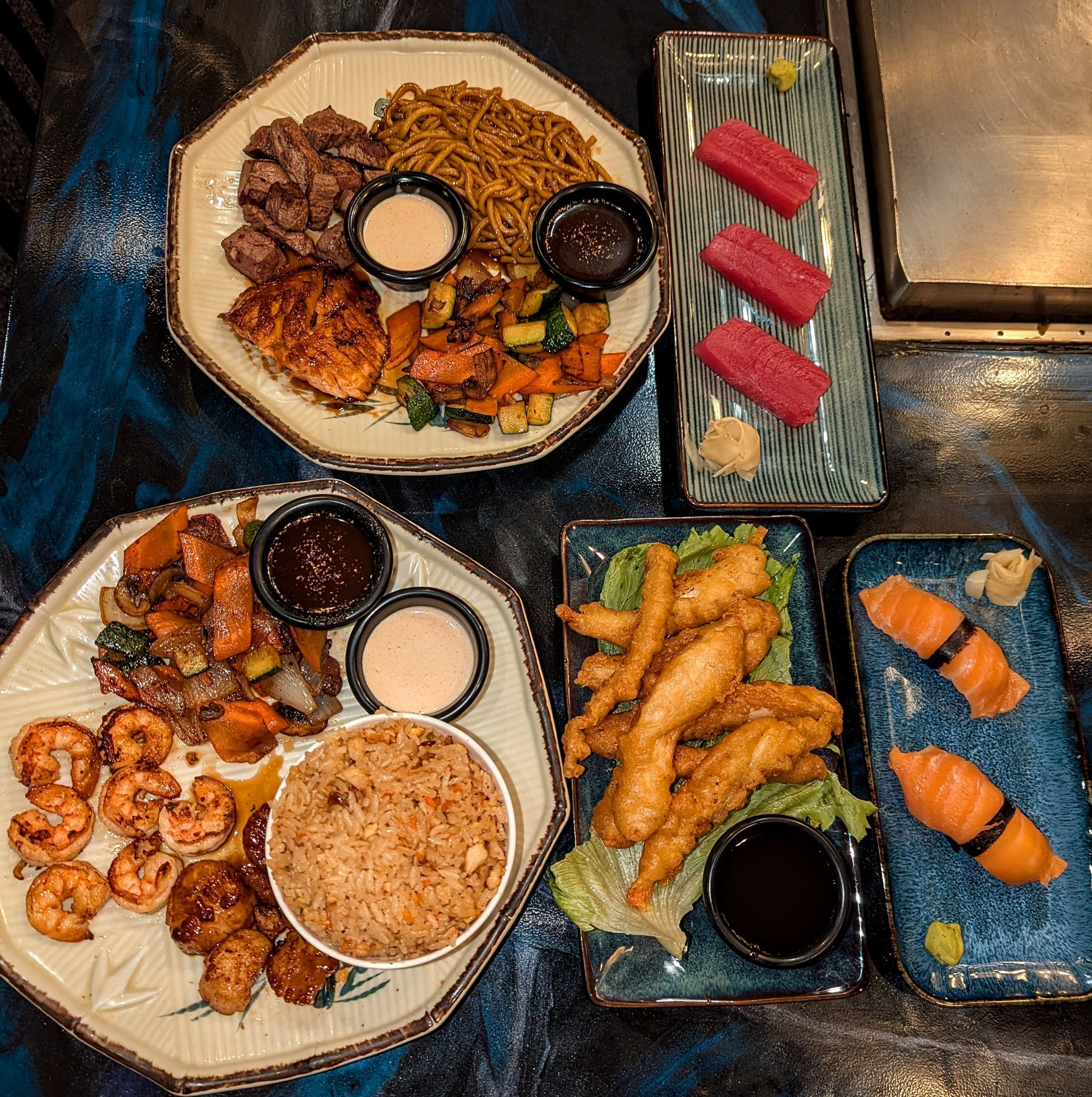 Assorted sushi and Asian appetizers on blue plates around dipping sauces, arranged on a table.