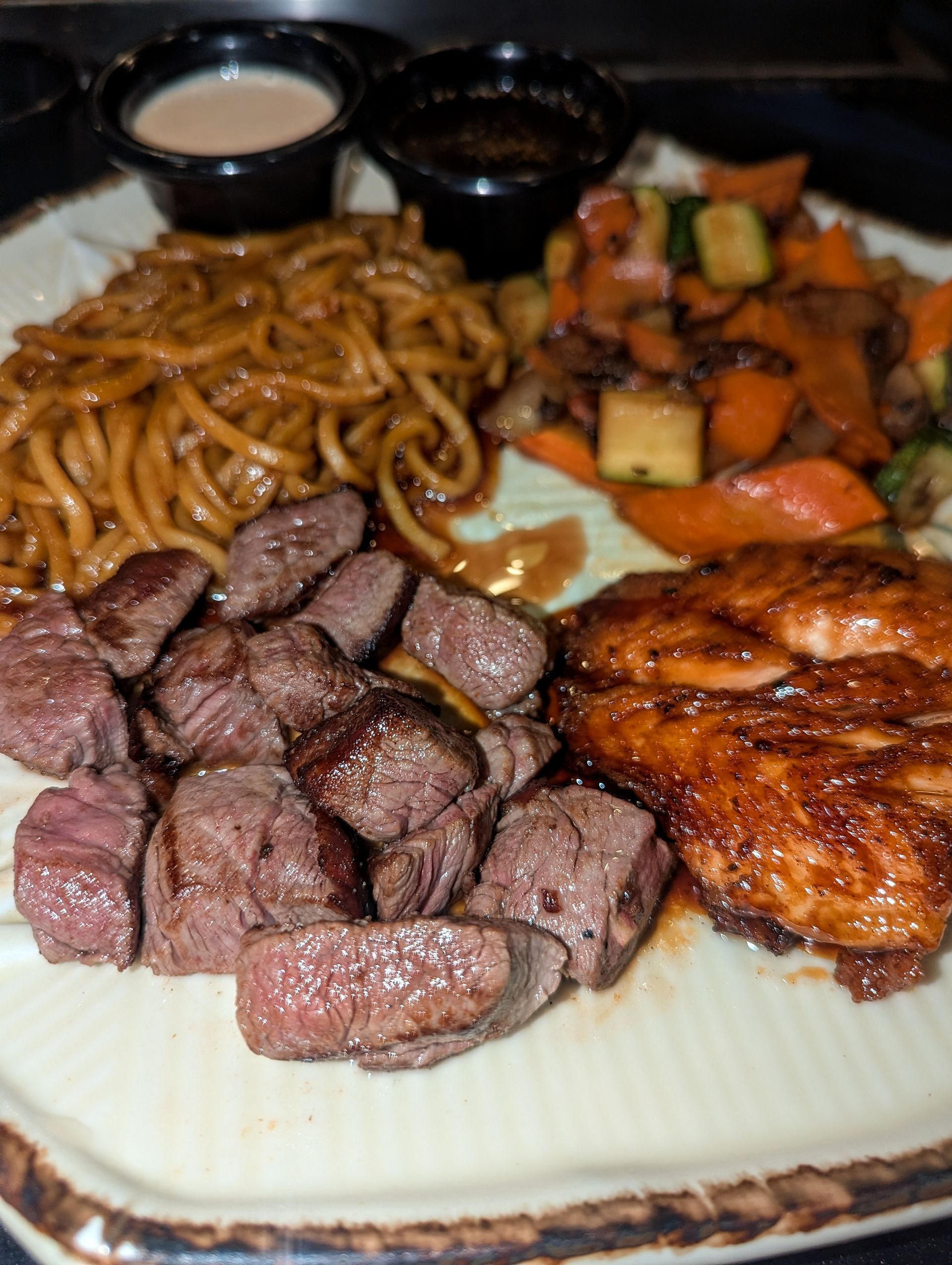 Steak, grilled chicken, noodles, and vegetables with dipping sauces on a plate