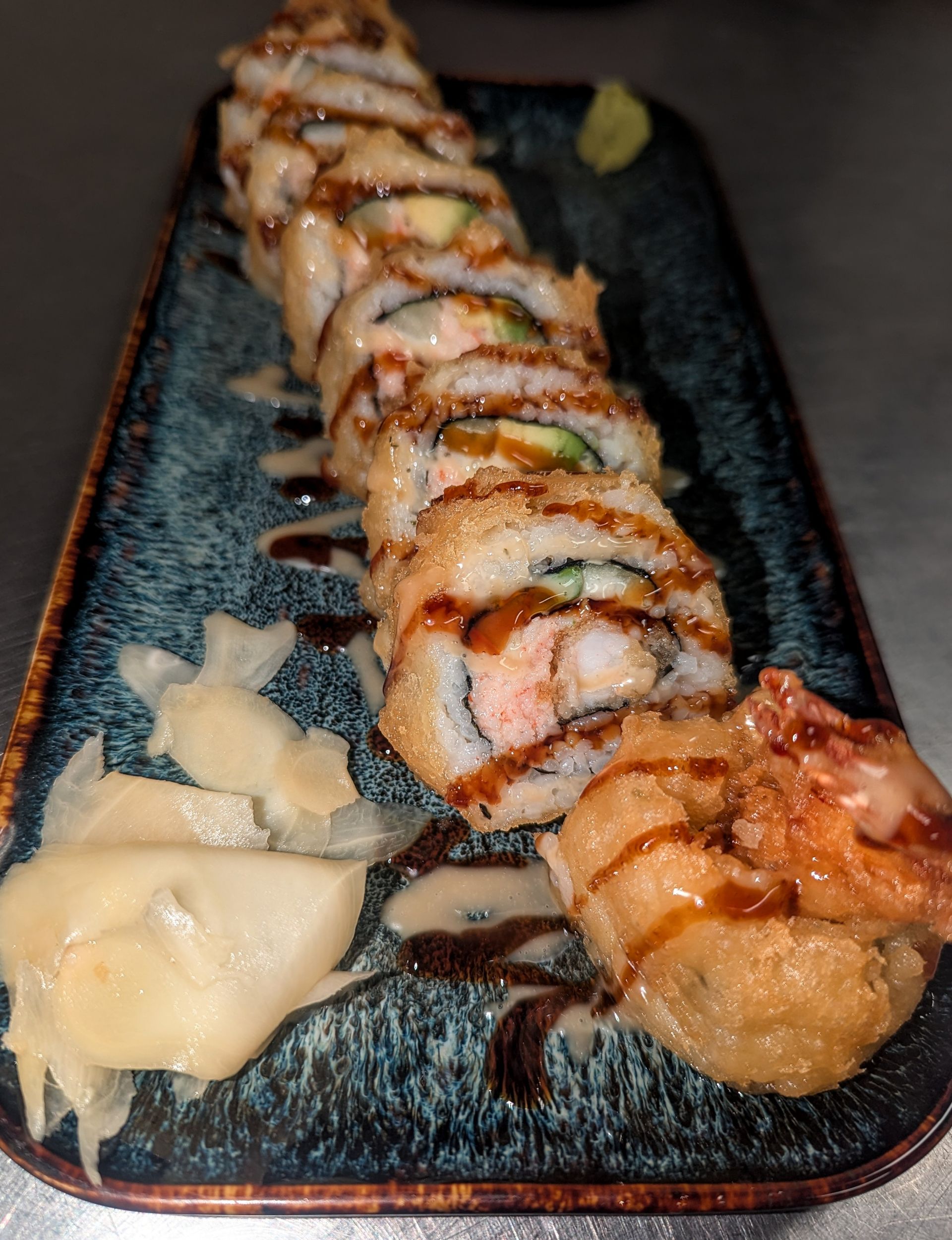 Sushi roll topped with sauce on a black plate, with pickled ginger and wasabi on the side