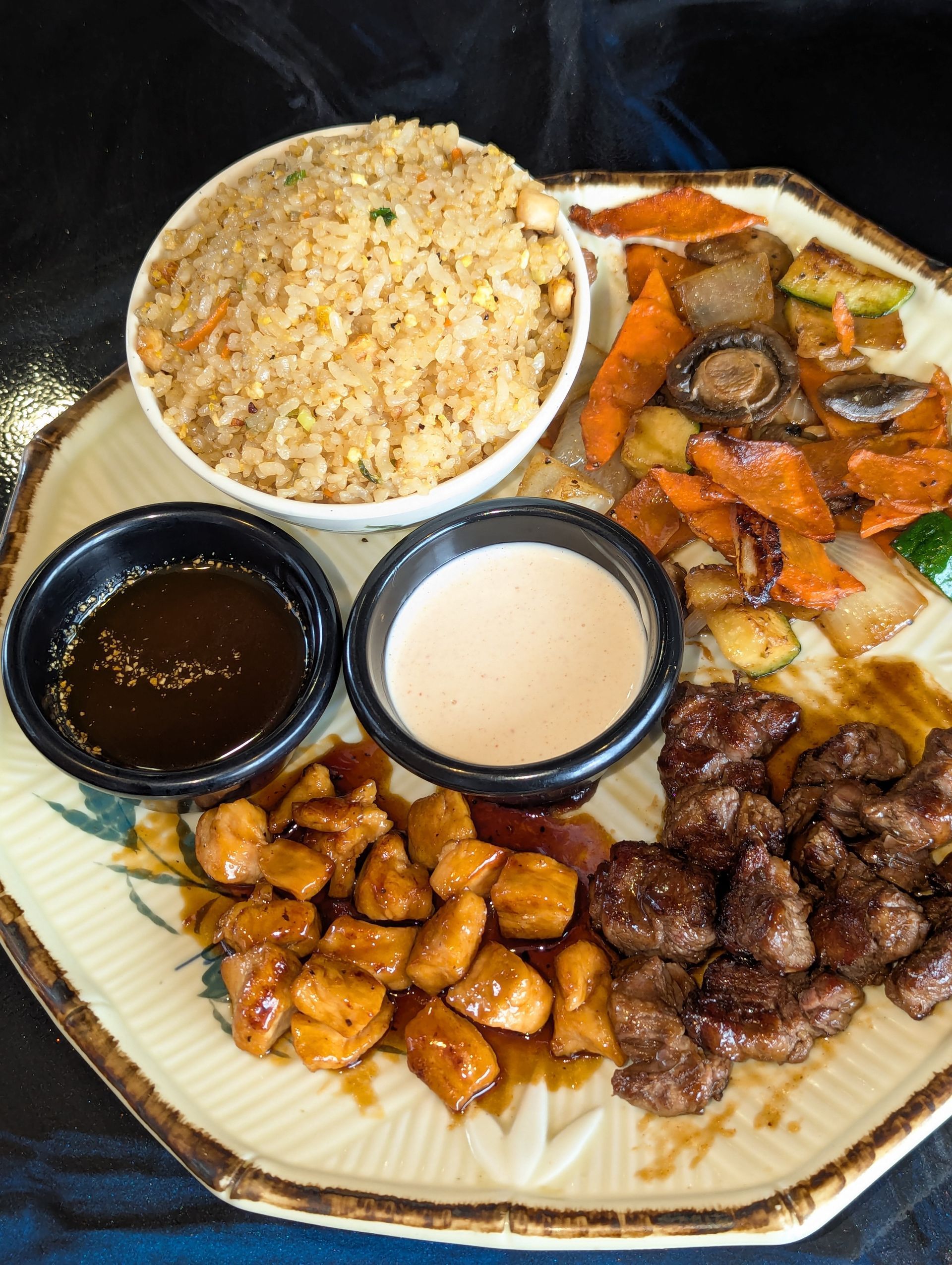 Plate with rice and assorted meats, vegetables, and two dipping sauces on a woven placemat.