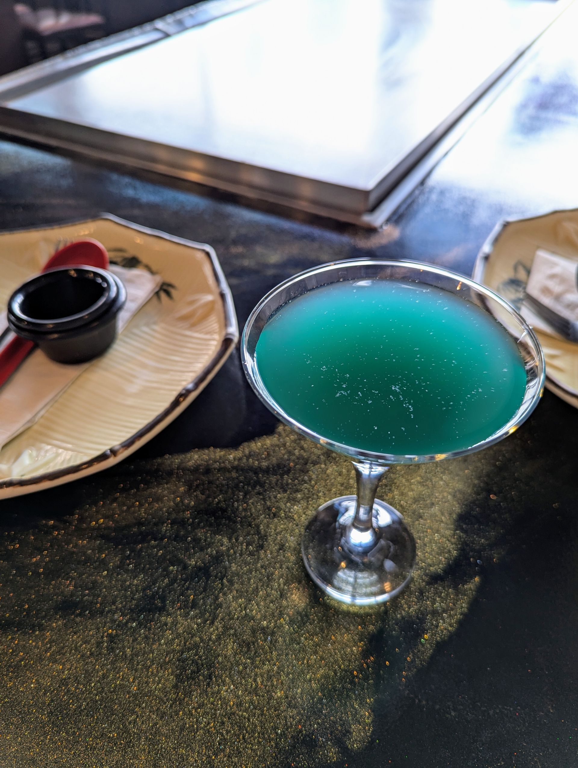 Two turquoise cocktails in martini glasses on a bar counter with small plates of snacks