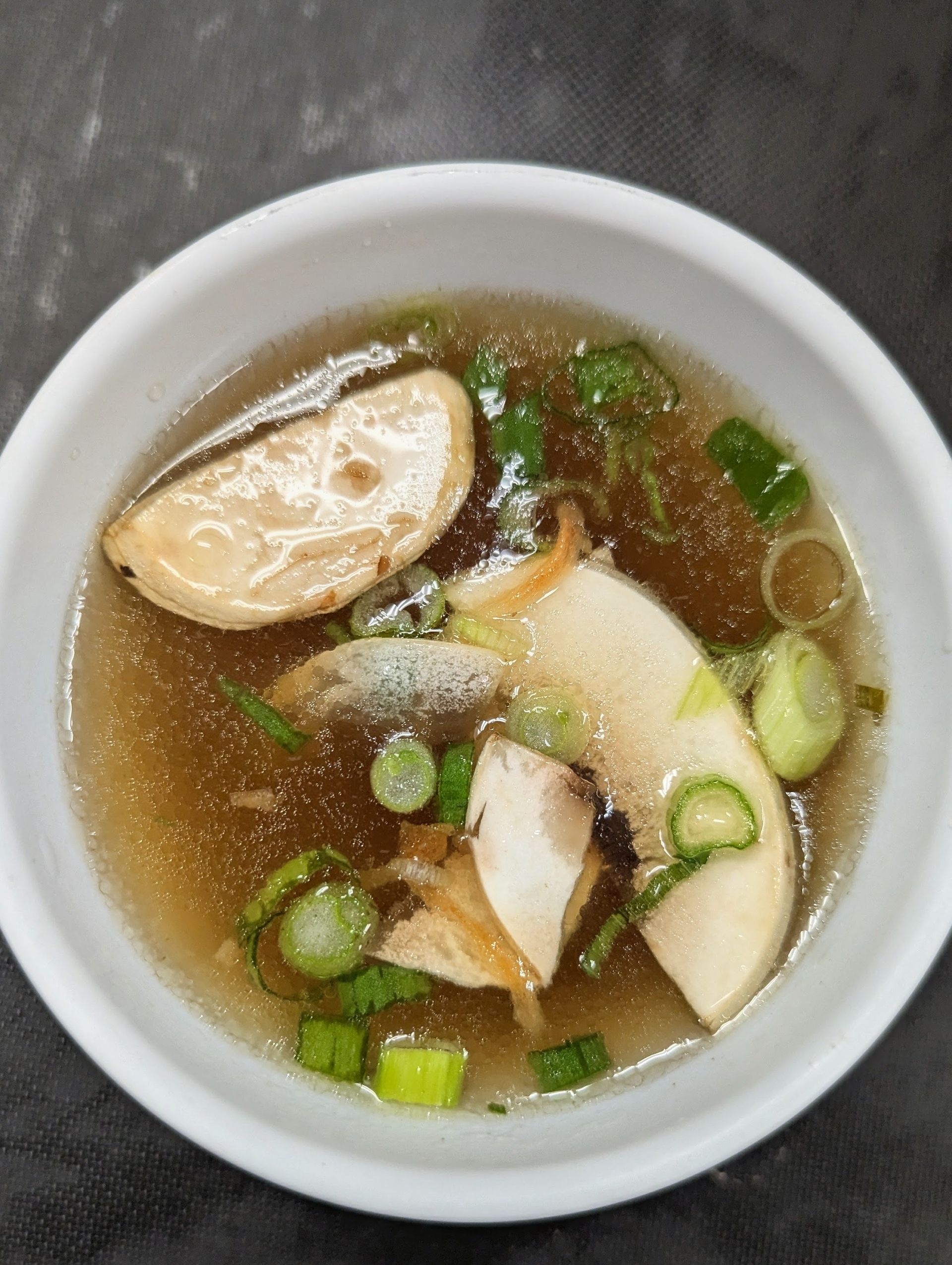 Bowl of clear soup with sliced fish cake and chopped green onions on a gray surface