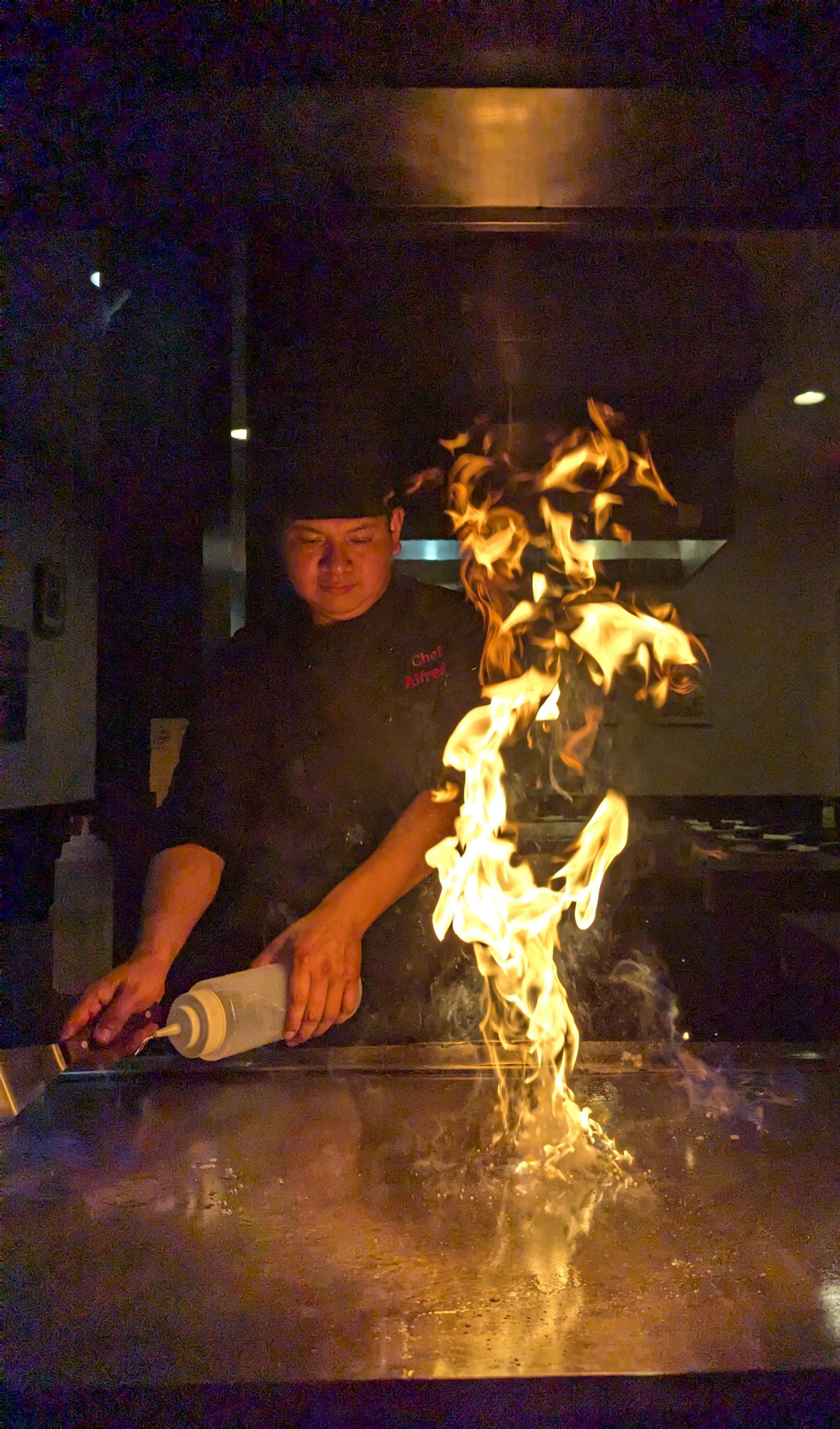 Chef performing a flaming tabletop cooking display in a dim restaurant.