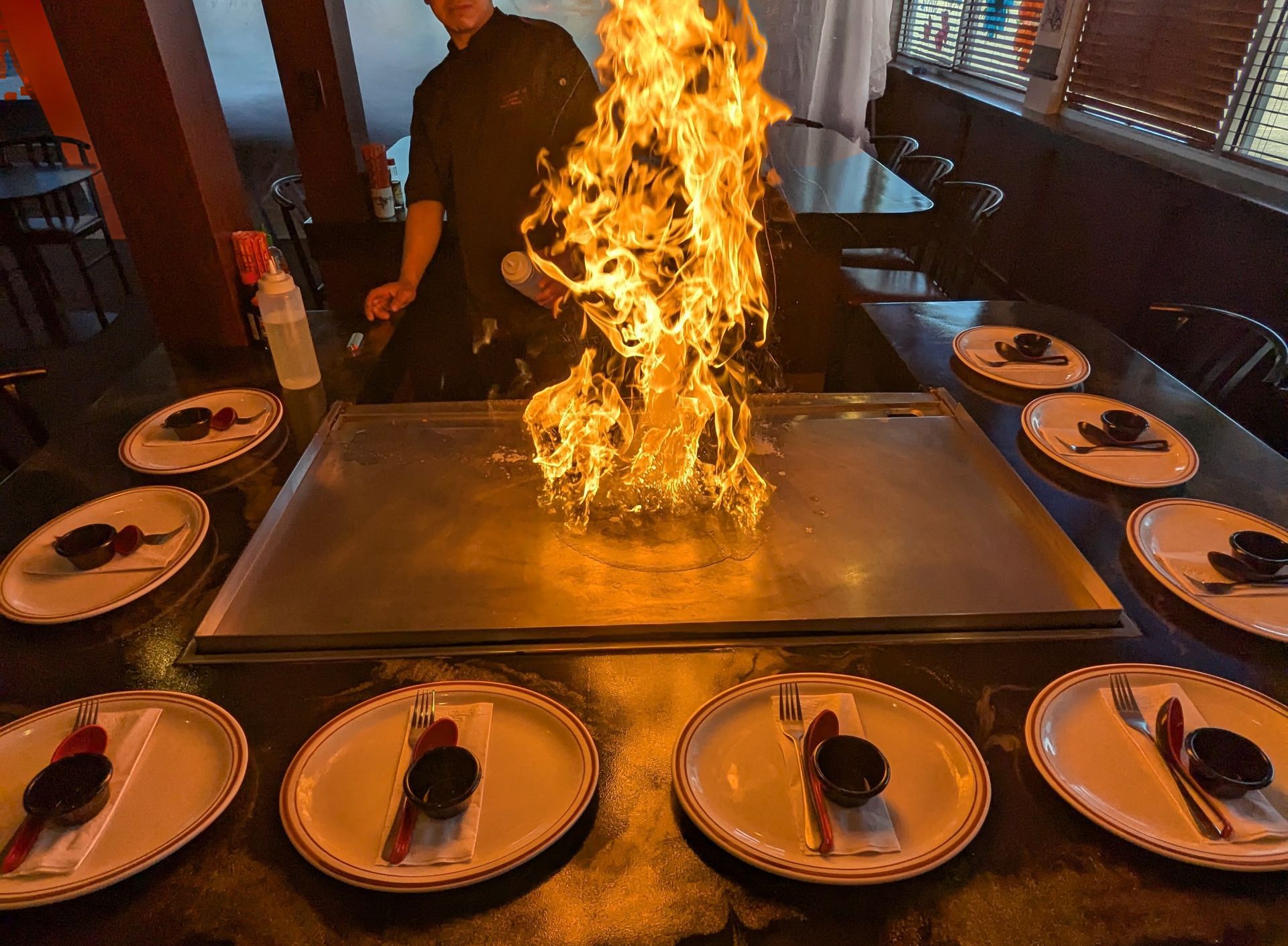 Restaurant teppanyaki grill with a chef igniting a large flame over the hot plate, plates set around it