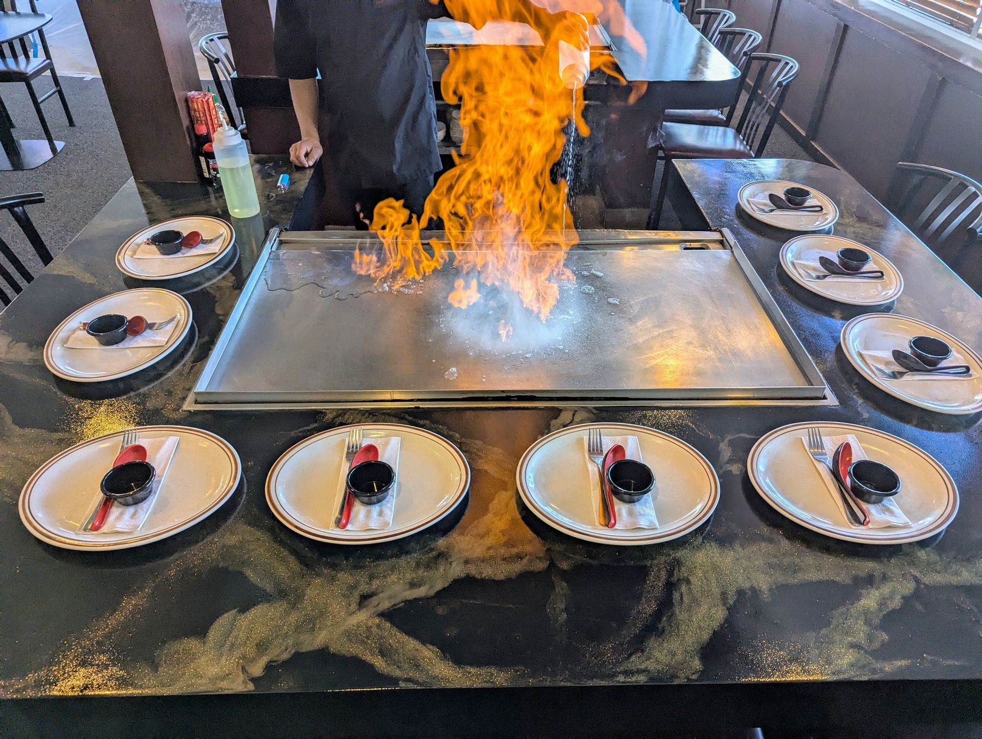 Flaming hibachi grill with chef cooking, plated dishes arranged around the table in a restaurant.