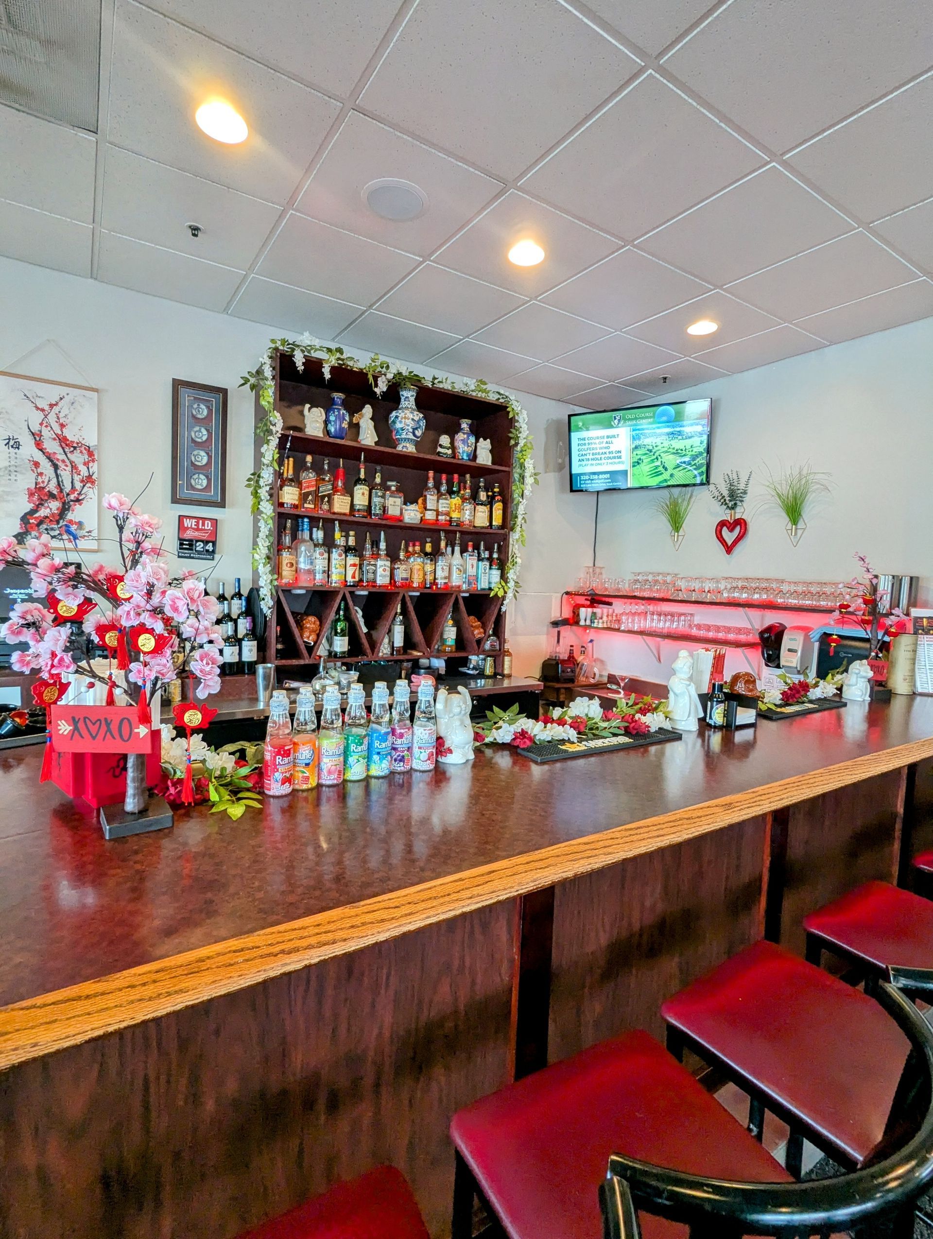Cozy bar interior with wooden counter, red stools, liquor shelves, and a TV on the wall.