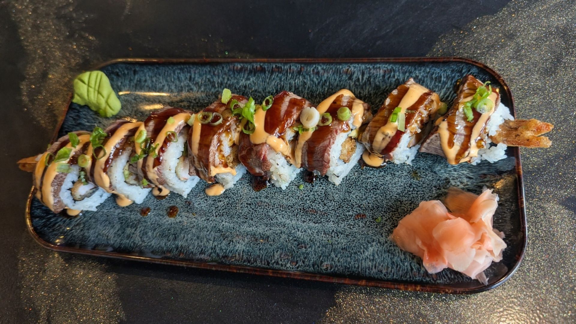 Sushi roll slices with sauce on a black rectangular plate, garnished with greens and pickled ginger.