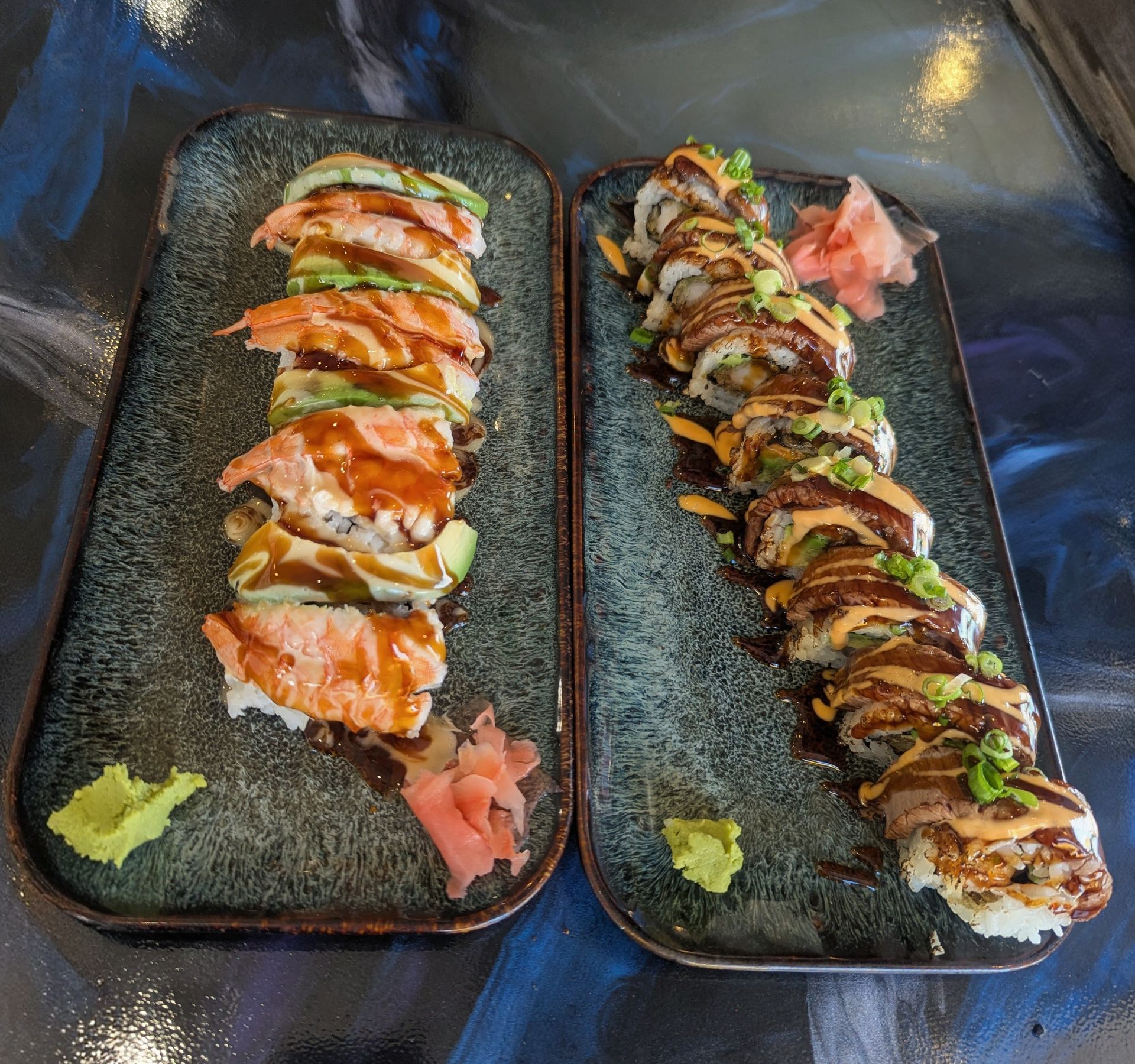 Two sushi platters with assorted rolls on black rectangular plates, with wasabi and pickled ginger
