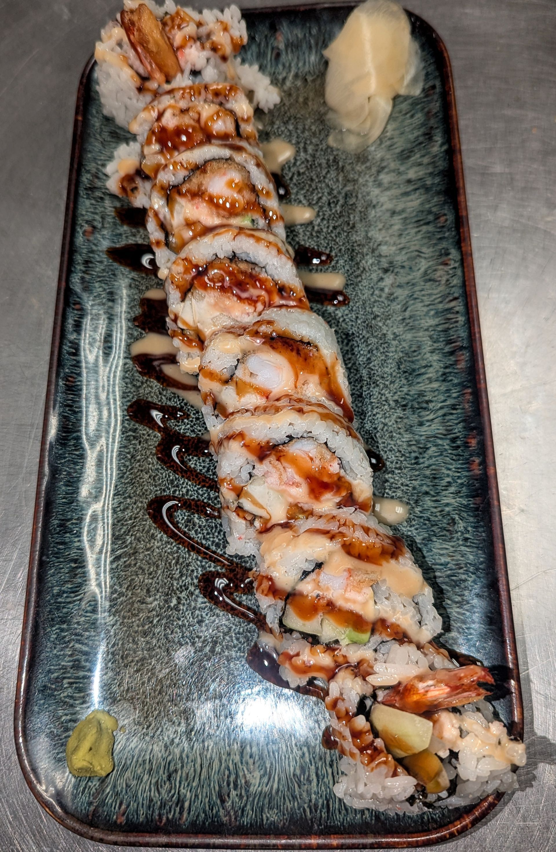 Sushi roll drizzled with sauce on a rectangular plate, with a small mound of wasabi on the side