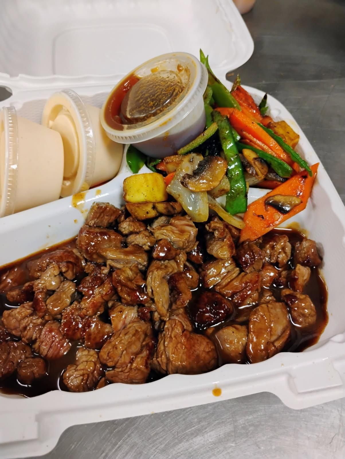 Takeout box with glazed meat, mixed vegetables, rice, and sauce cup