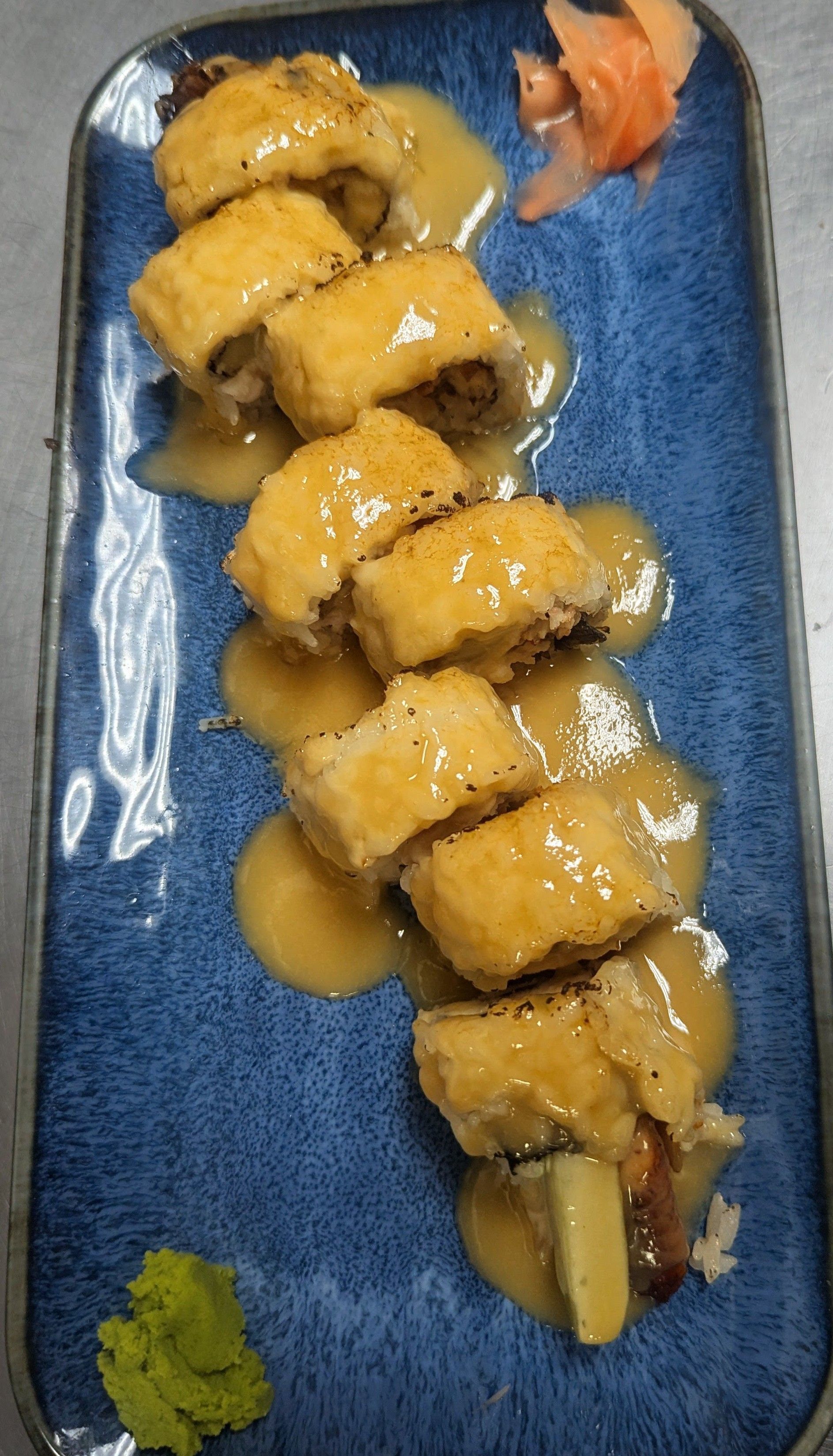 Blue plate with sushi rolls drizzled in sauce, garnished with wasabi and pickled ginger.