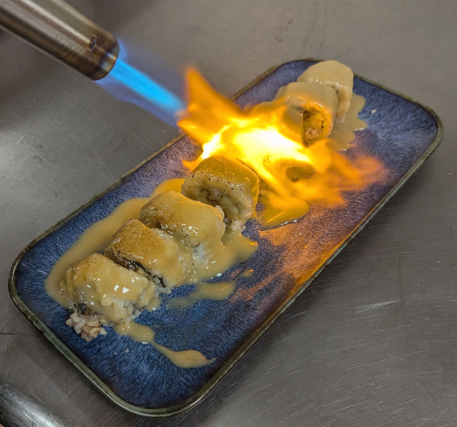Blue-flamed torch melting food on a blue plate, with bright orange flames and melted cheese spilling over the edges