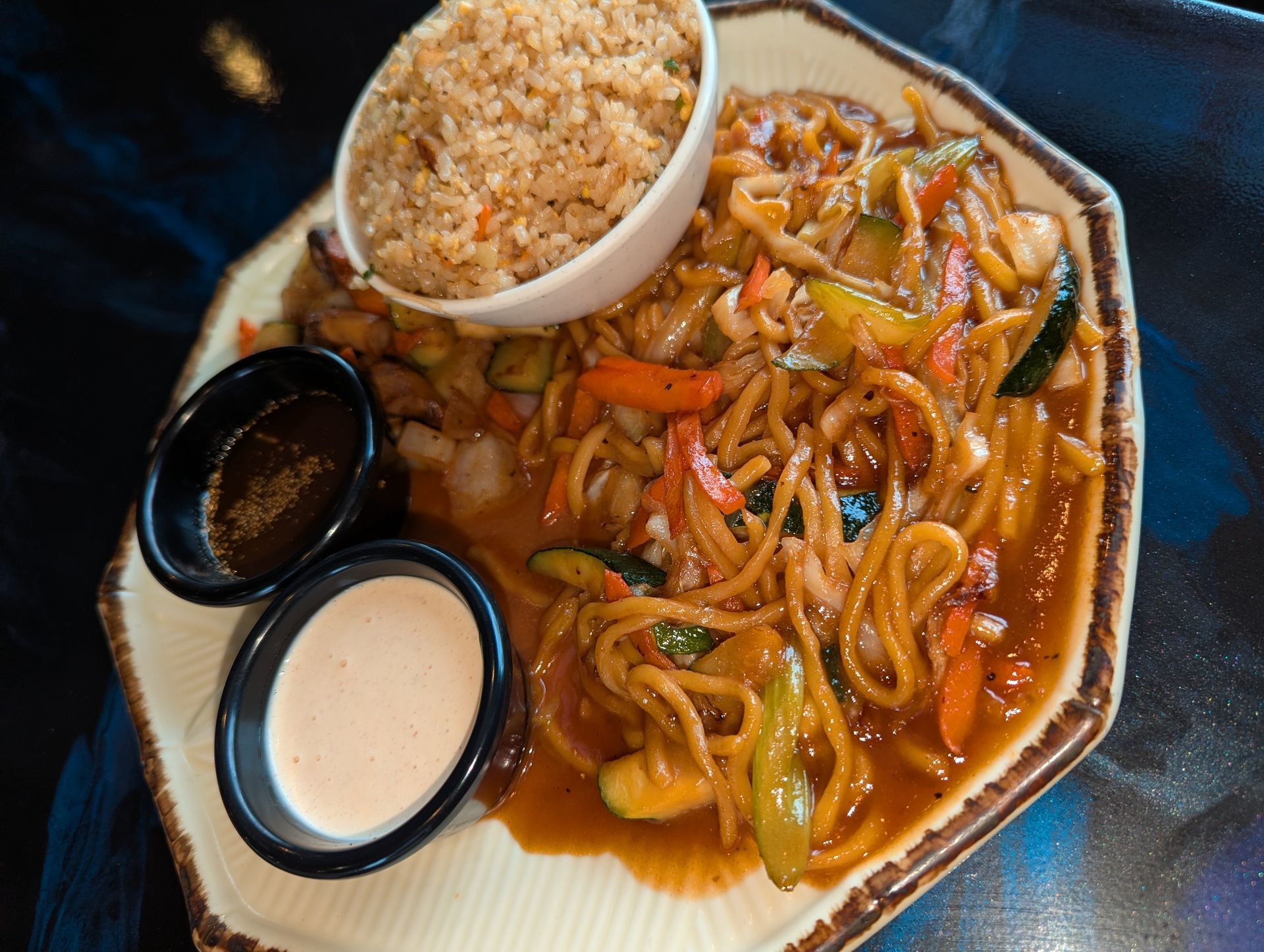 Plate of noodles with vegetables, fried rice, and two dipping sauces on a dark table