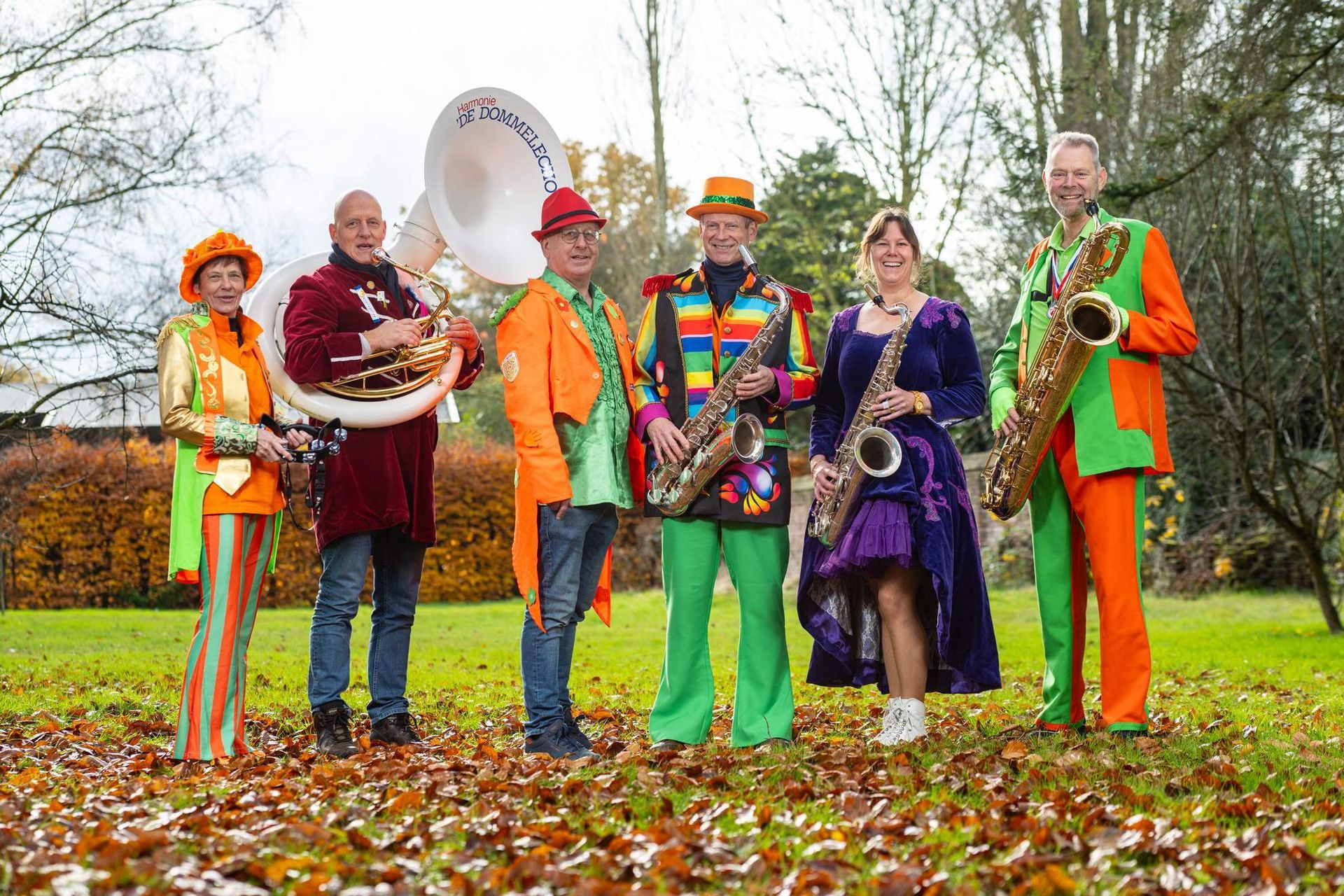 Een muzikaal ensemble in kleurrijke kostuums poseert buiten in de herfstbladeren.
