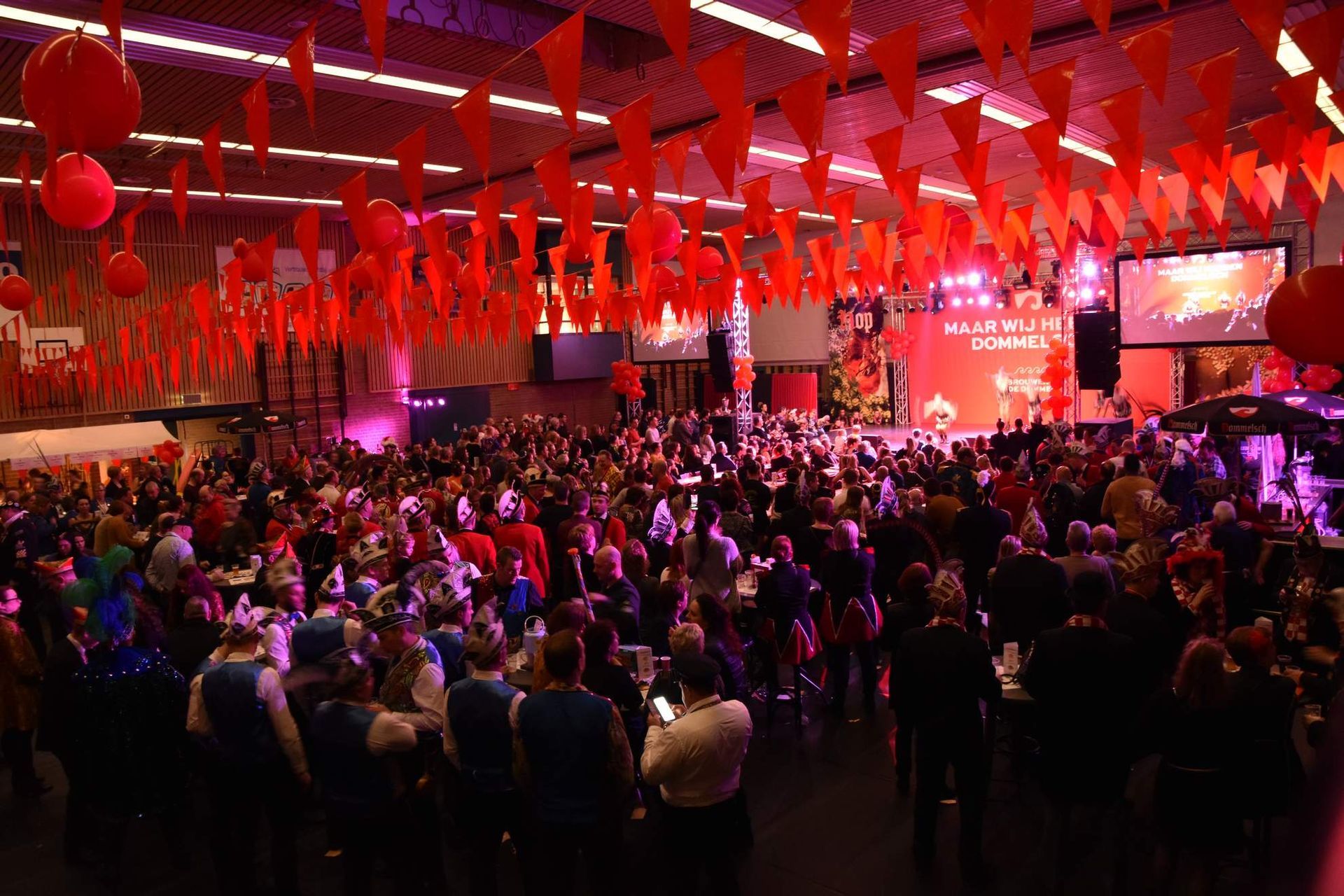 Grote menigte bij een evenement, rode versieringen aan het plafond, podium met scherm zichtbaar.