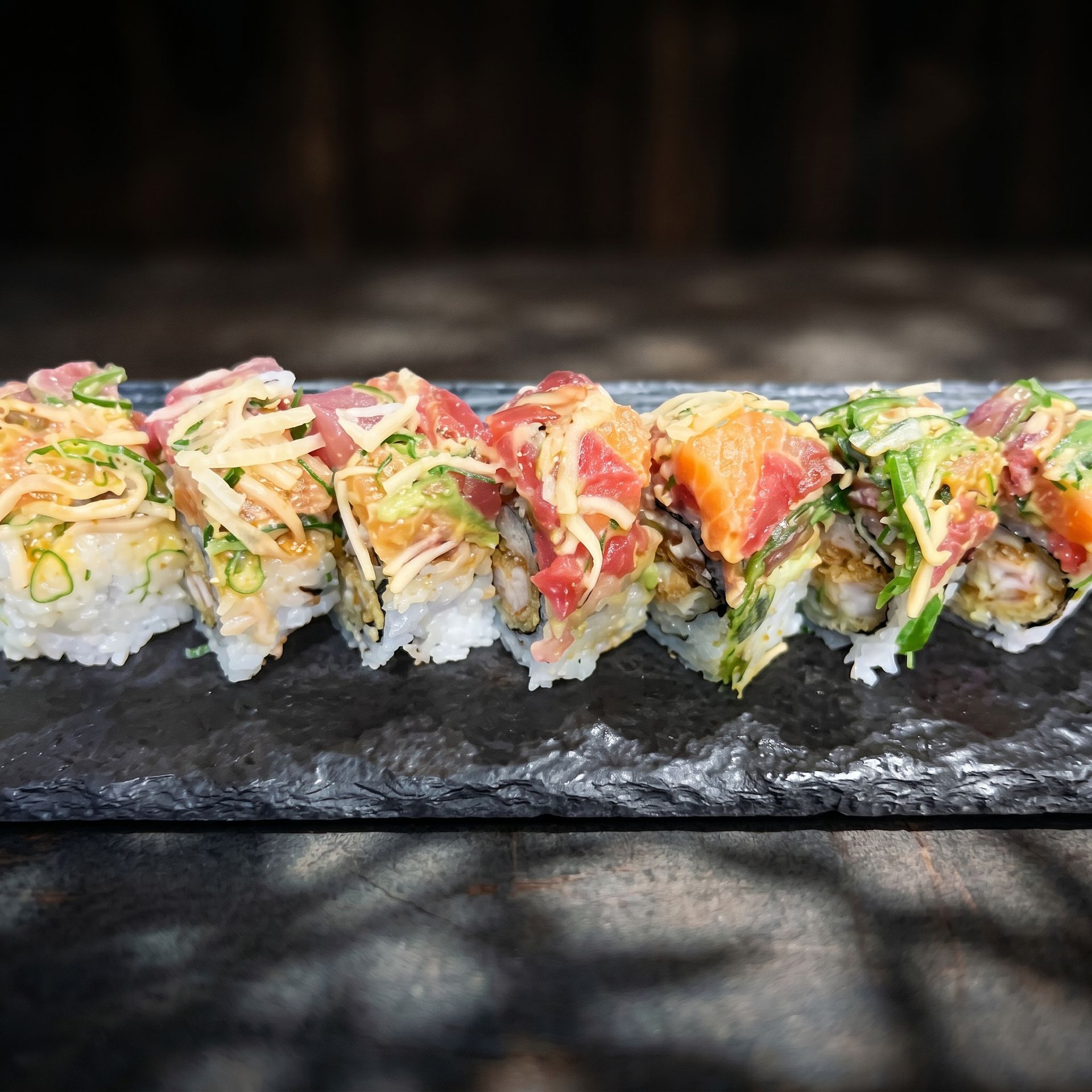 Colorful sushi rolls topped with avocado, fish, and sauces on a dark slate platter