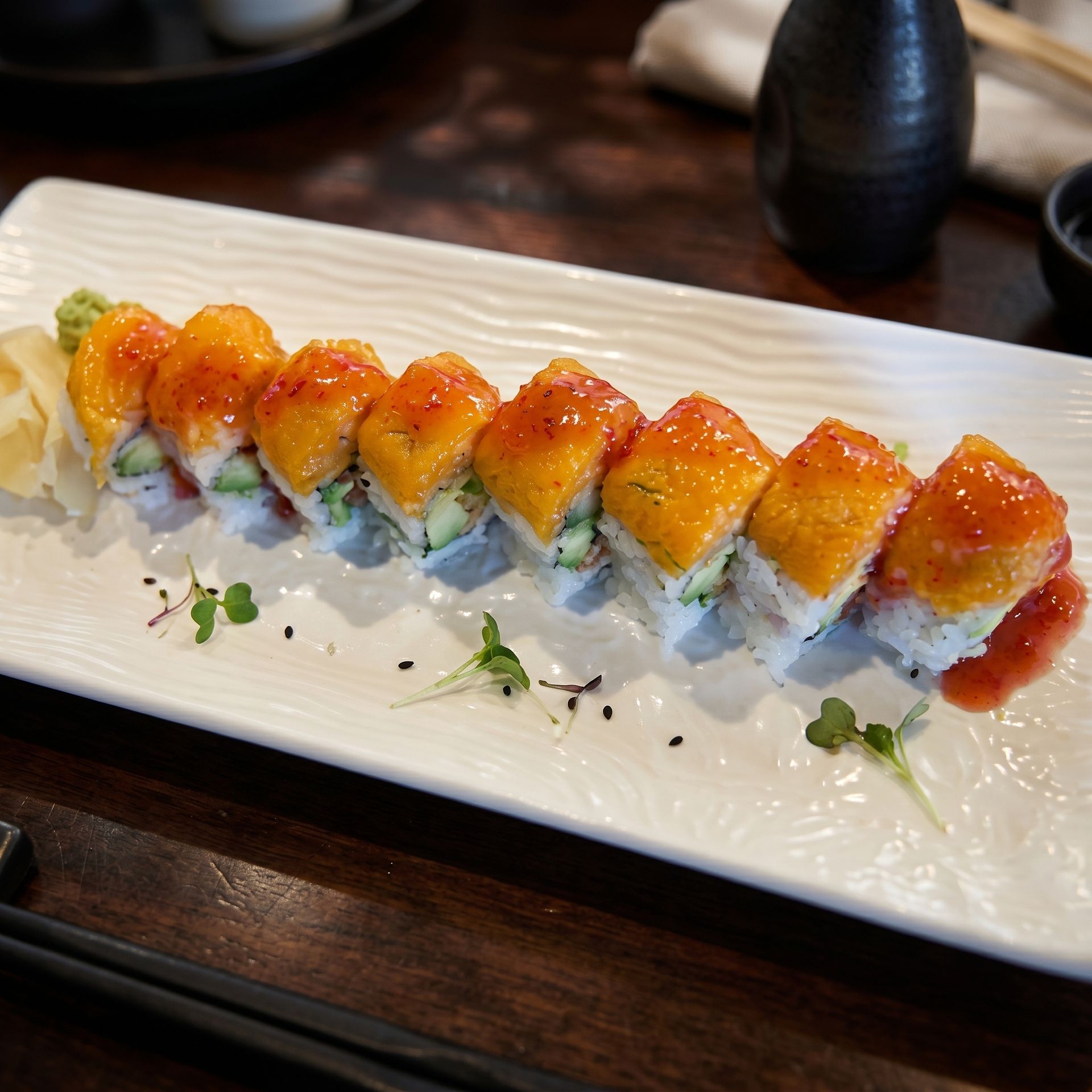 Sushi roll topped with golden sauce on a white rectangular plate, garnished with microgreens