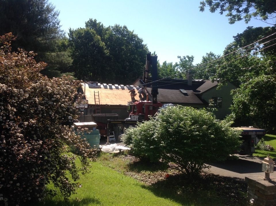 House with roofing being replaced, workers on the roof, surrounded by trees and lush green bushes on a sunny day.