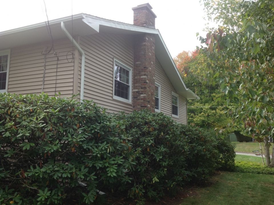 Tan house with brick chimney and green bushes in front. Windows and trees visible.
