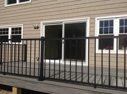 A deck with black metal railings in front of a house with a sliding glass door and windows; siding is tan.
