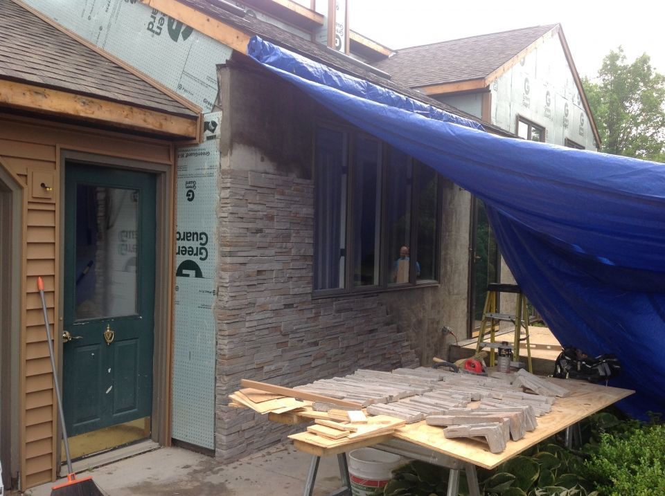 Exterior house under construction, stone facade being installed. Blue tarp covers side wall and construction materials sit on plywood table.