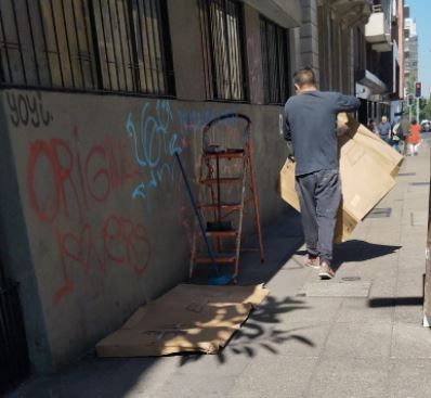 Persona llevando cartón por una acera de la ciudad, junto a una pared con grafitis y carros apilados