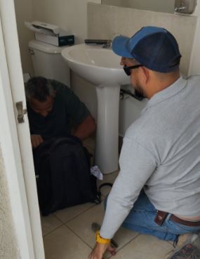 Dos personas están agachadas junto al lavabo de un baño, inspeccionando algo cerca de una bolsa negra en el suelo.