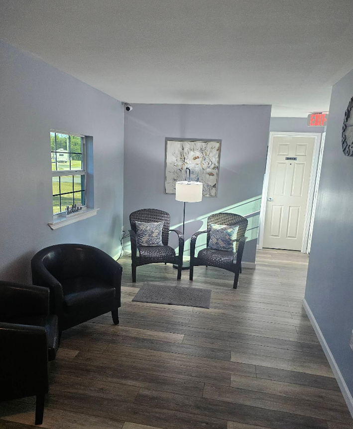 A waiting area with two patterned chairs, a black chair, and a small window. Pale blue walls, wood floor.