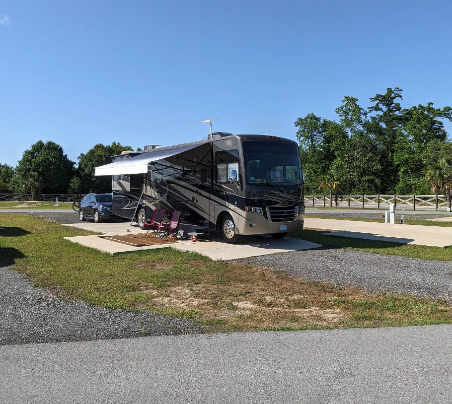 RV parked at campsite with awning extended, parked car, trees, blue sky.