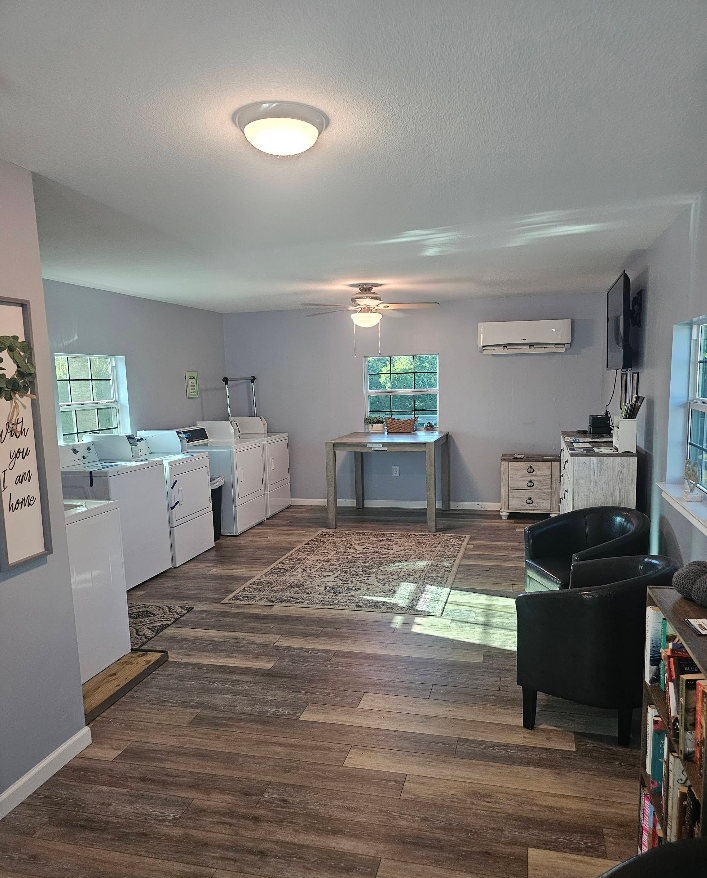 Laundromat interior with washers, dryers, seating area, and table. Gray walls, wood-look flooring, and a rug.
