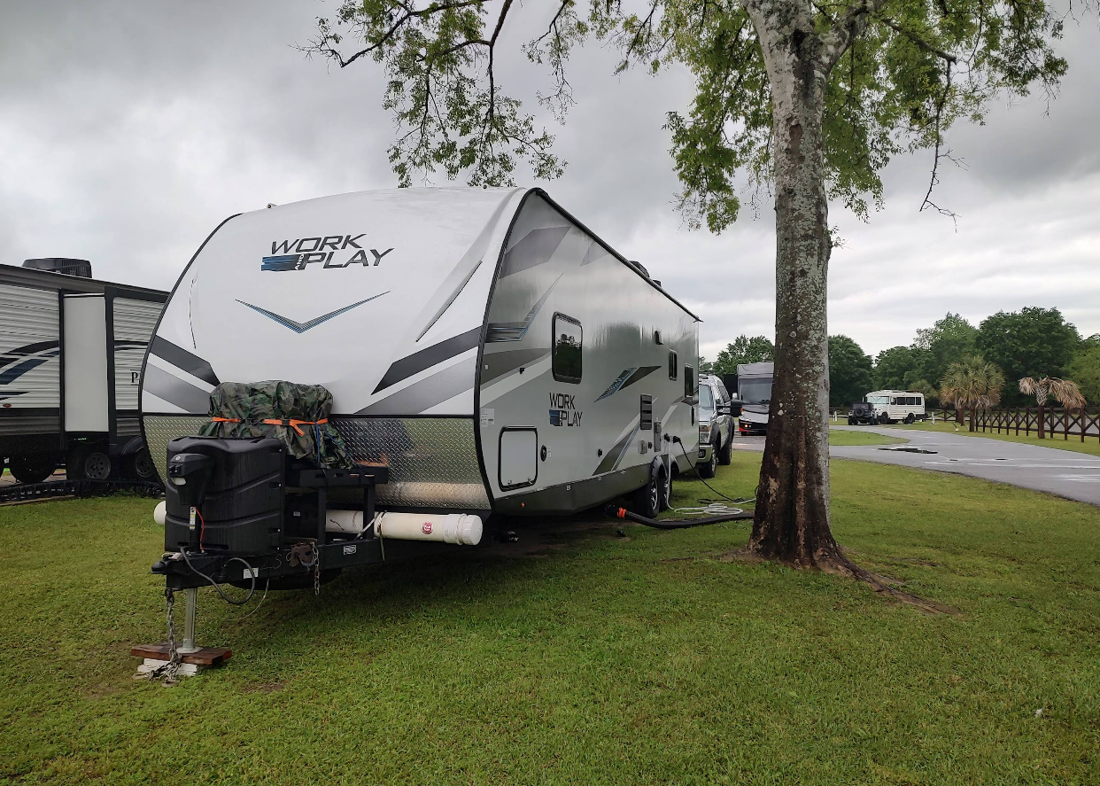 A parked travel trailer on grass beside a tree, in a campground on an overcast day.