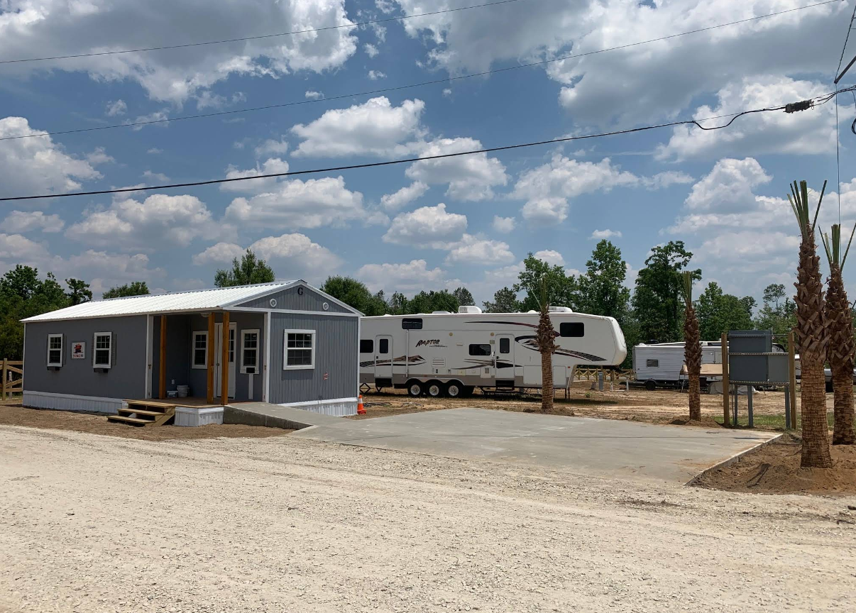Campsite with a gray cabin, RVs, gravel, palm trees, and a cloudy sky.
