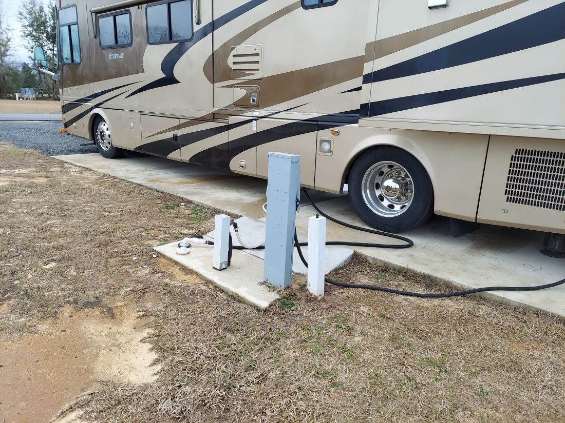 RV parked at campsite; electrical and water hookups visible. Beige and brown RV, concrete pad.