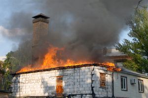 House roof engulfed in flames with thick gray smoke rising above a chimney