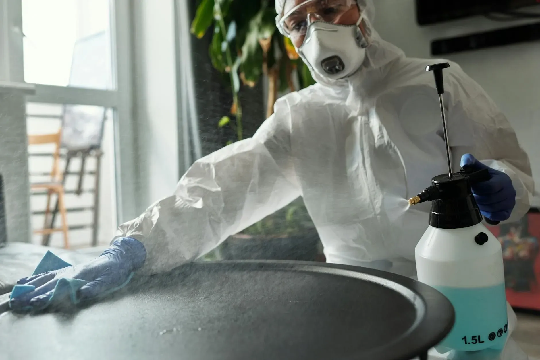 Person in protective gear disinfecting a table with a spray bottle and cloth inside a bright room