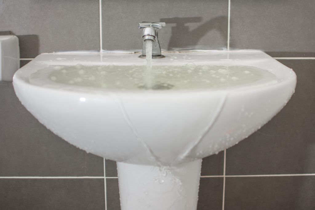 White bathroom sink with running faucet and water overflow staining the basin and pedestal.